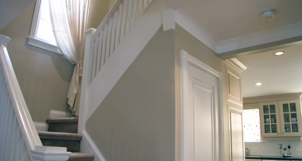 A view of a staircase with white railing, a small window, and a door, next to a kitchen with white cabinets.