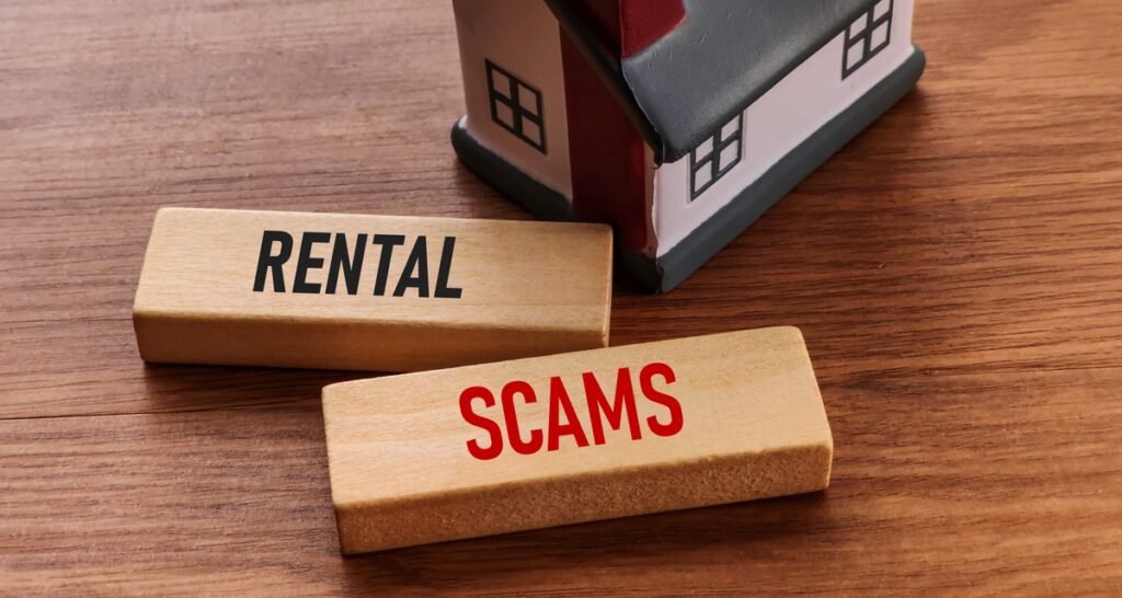 Two small rectangular wood blocks displaying the words “rental” and “scams” placed on a desk next to a toy house.
