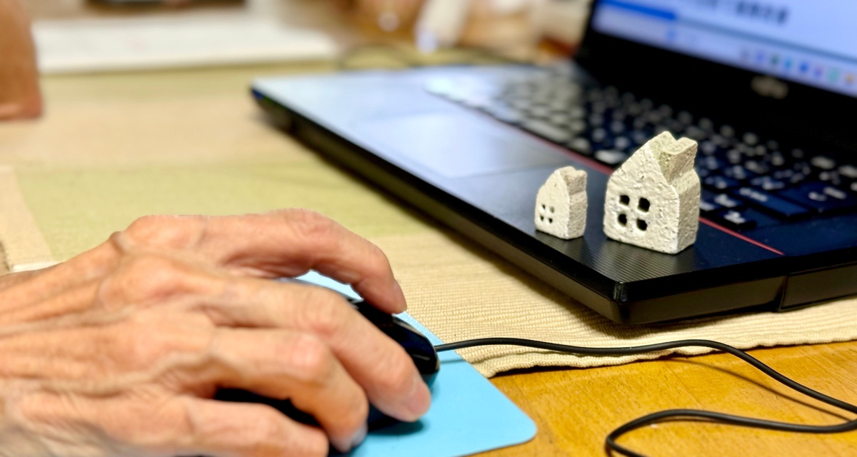 A close-up of a hand holding a computer mouse next to a laptop on a desk with two small white clay houses.