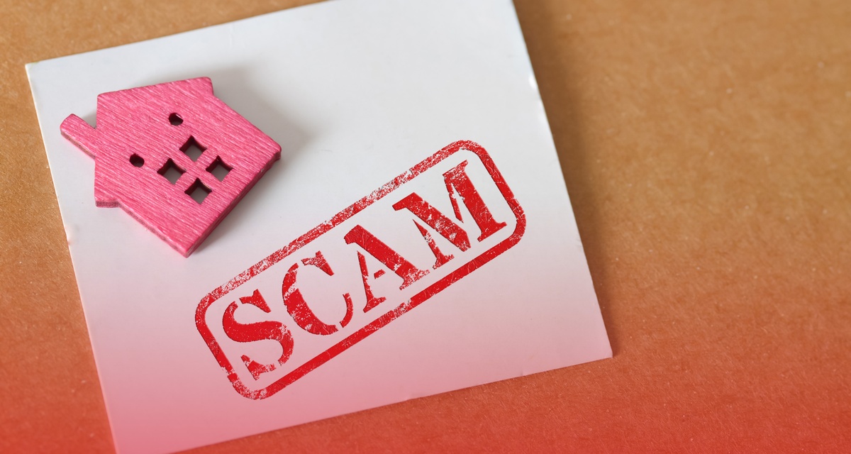 An aerial view of a white sheet of paper displaying the word “scam” next to a small red wooden house on a desk.