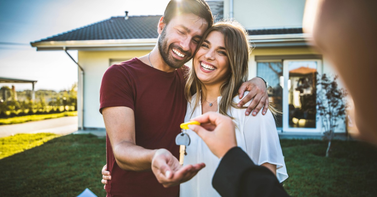 A man holding a woman while they both smile, with a person handing them a set of keys with a house in the background.