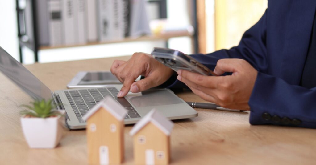 A close-up of the hands of a person holding a smartphone while using a laptop on a desk with small wooden houses.