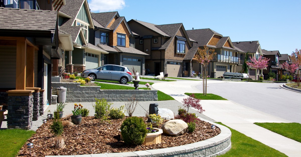 Multiple two-story homes in a row with front lawns and driveways next to a concrete street on a sunny day.