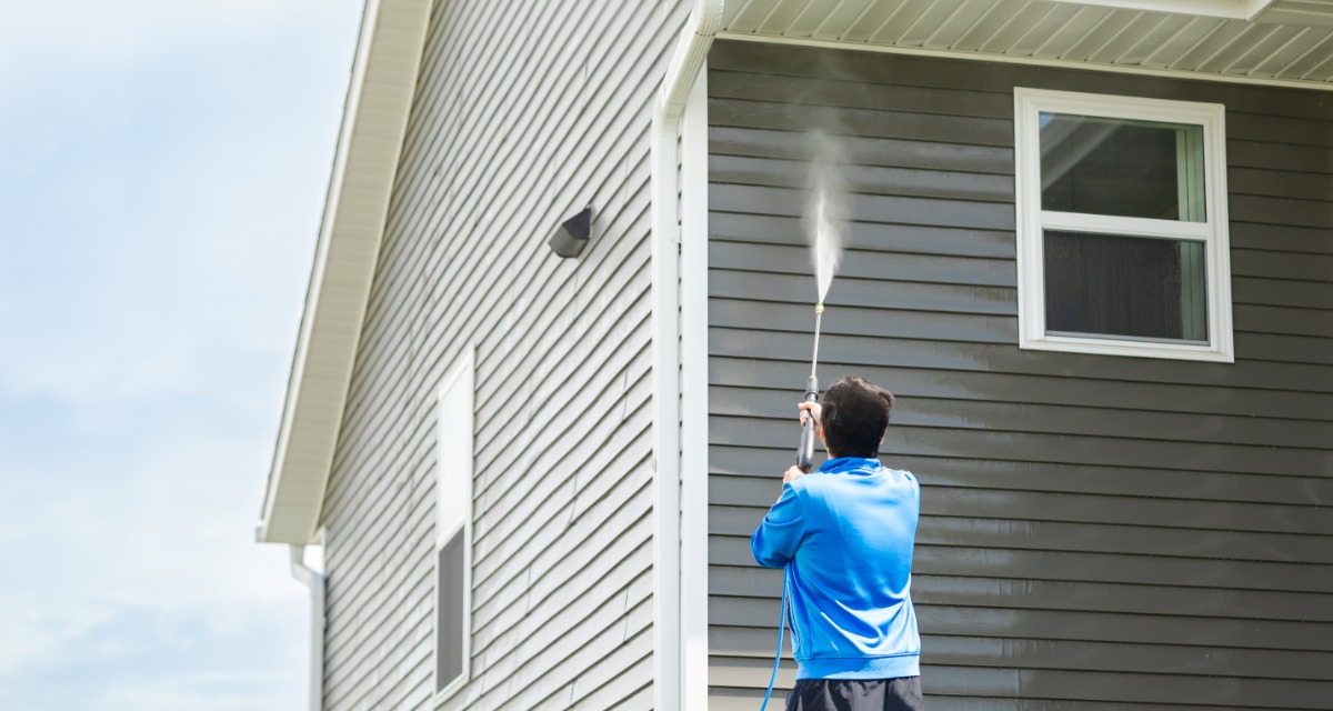 A person wearing a blue sweatshirt using a pressure washer to clean the exteriors of a two-story home on a sunny day.
