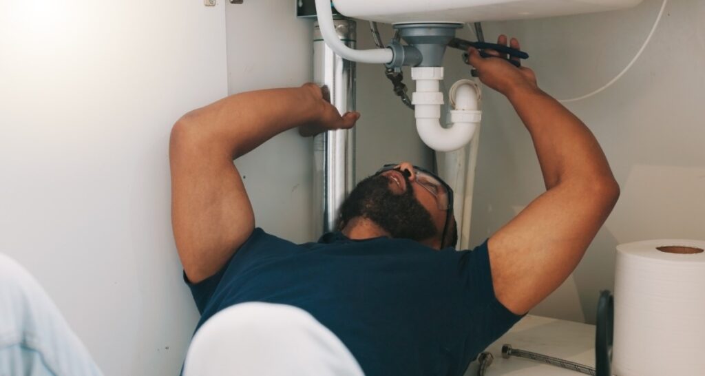 A worker wearing a polo shirt and protection glasses, holding a small wrench next to a tube under a white sink.