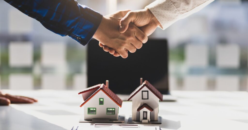 A close-up of two hands holding on top of a white desk with a laptop and two model homes with small chimneys.