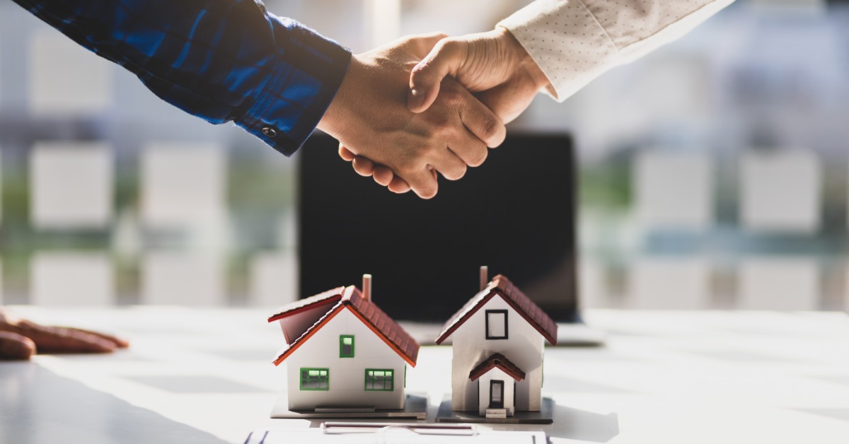 A close-up of two hands holding on top of a white desk with a laptop and two model homes with small chimneys.