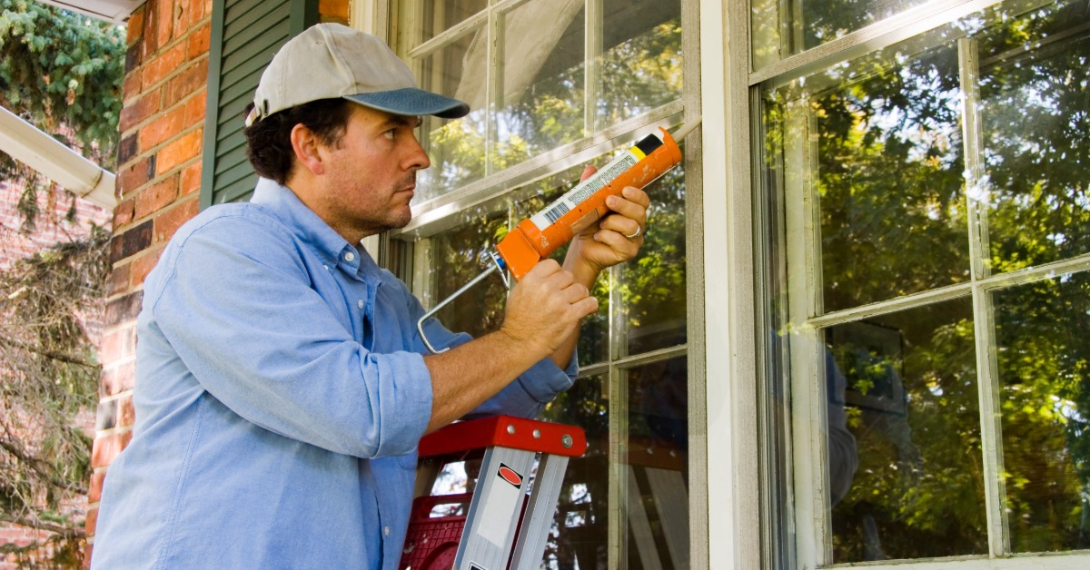 A man wearing a blue shirt and a hat standing on a stainless steel ladder while using a caulk gun to seal a window.