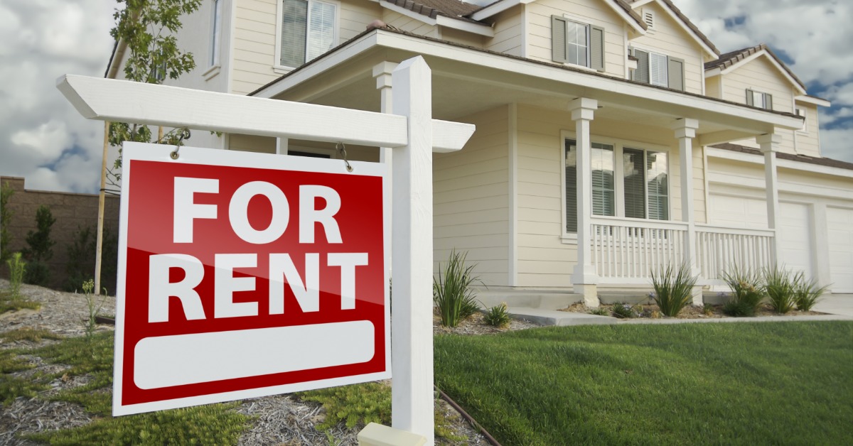A close-up of a red “for rent” sign on a white structure, placed in front of a two-story home with green grass.