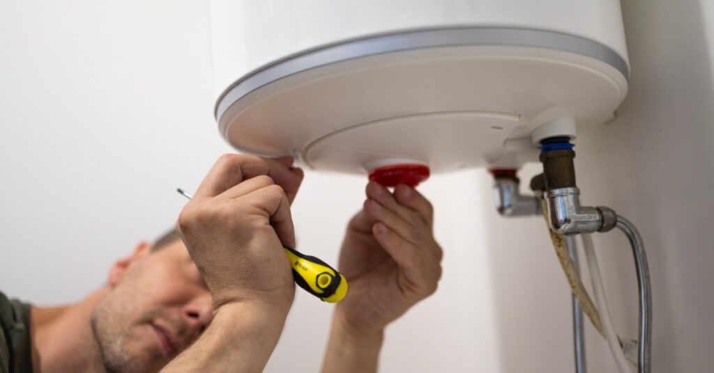 A man holding a screwdriver while touching the underpart of a water heater placed on the wall inside a room.