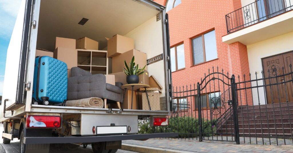 A large moving truck with the back doors open, showing furniture and boxes, parked outside a brick townhome.