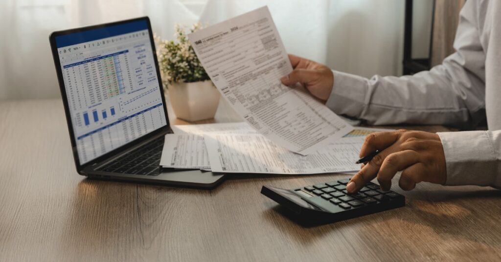 A close-up of the hands of a man holding a piece of paper while using a calculator on a desk next to an open laptop.