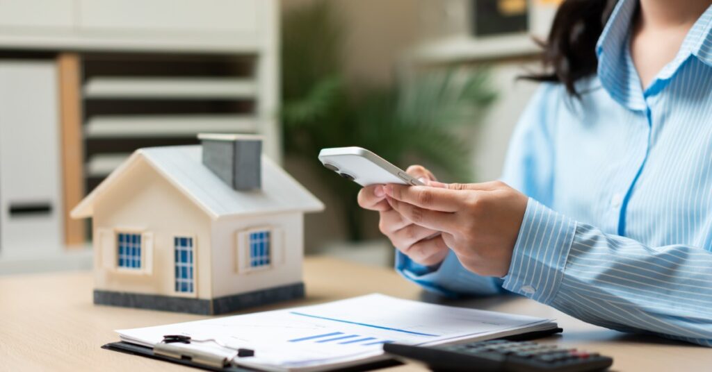 A close-up of the hands of a woman using a smartphone, sitting behind a desk with a small model house and documents.