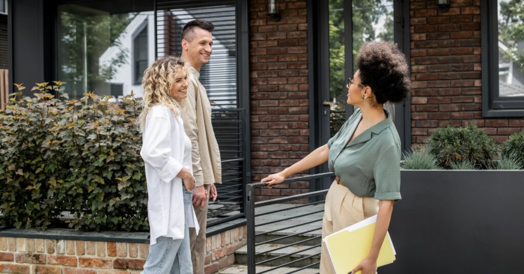 A woman holding a folder while holding on to a metal rail and talking with a couple outside of a brick home.