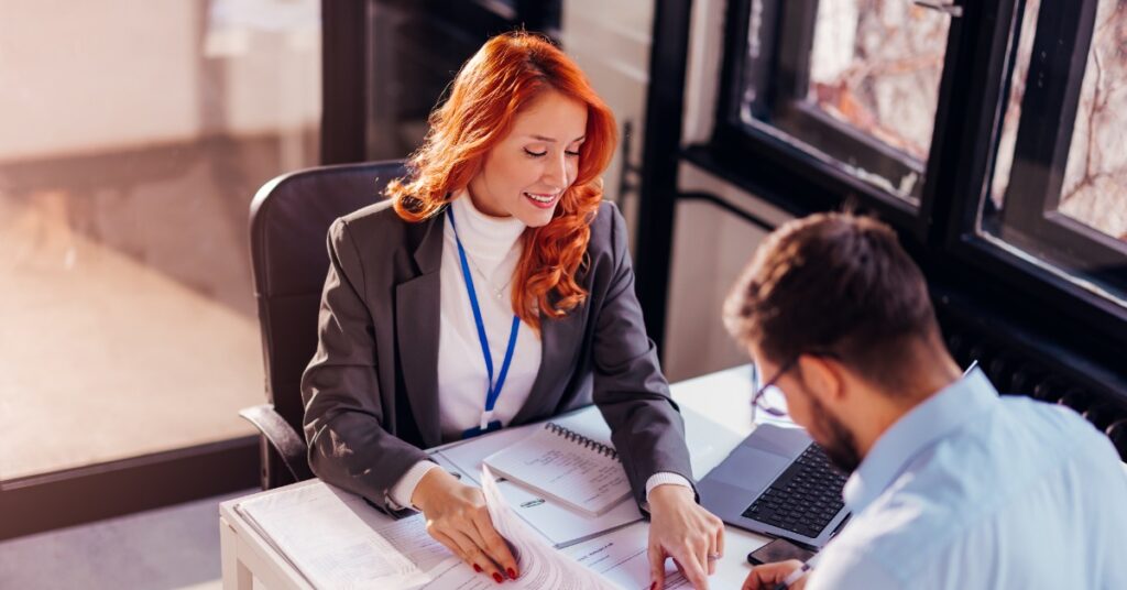 A woman sitting behind a desk wearing a lanyard, pointing at a document for a man sitting in front of her to sign.