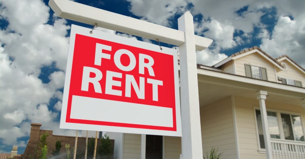 A close-up of a red “For Rent” sign with a white holder placed in the front of a two-story home with clouds in the sky.