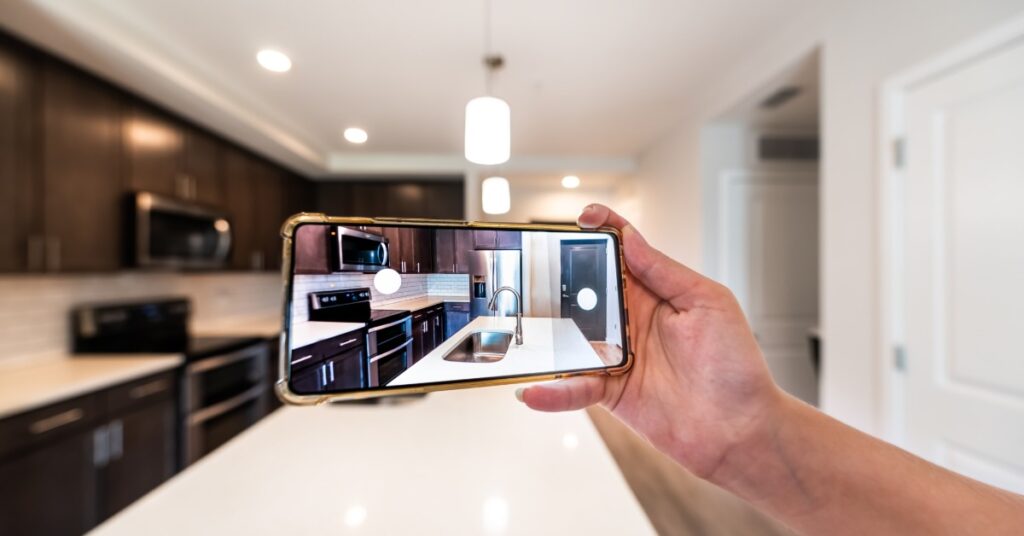 A close-up of a hand holding a smartphone, taking a picture of a home’s kitchen with a white counter and brown cabinets.