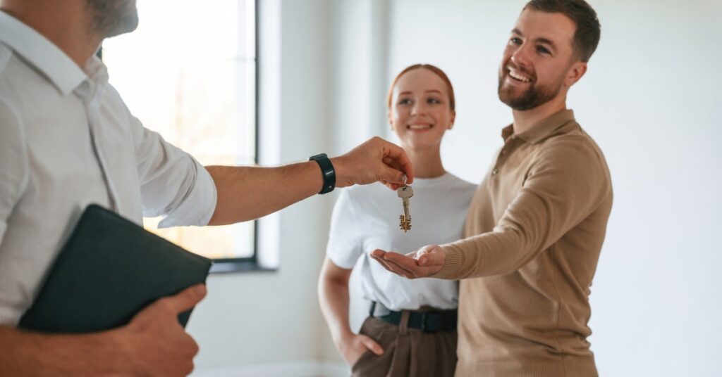A man wearing a white shirt holding a binder while handing a set of keys to a couple standing in an empty room.