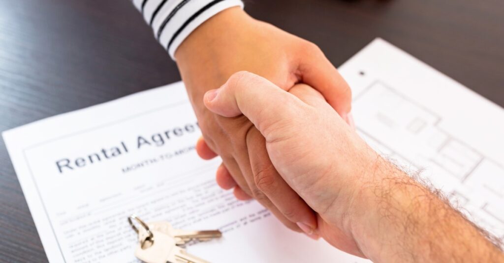 A close-up of two shaking hands over a desk with a paper displaying the words “rental agreement” next to a set of keys.