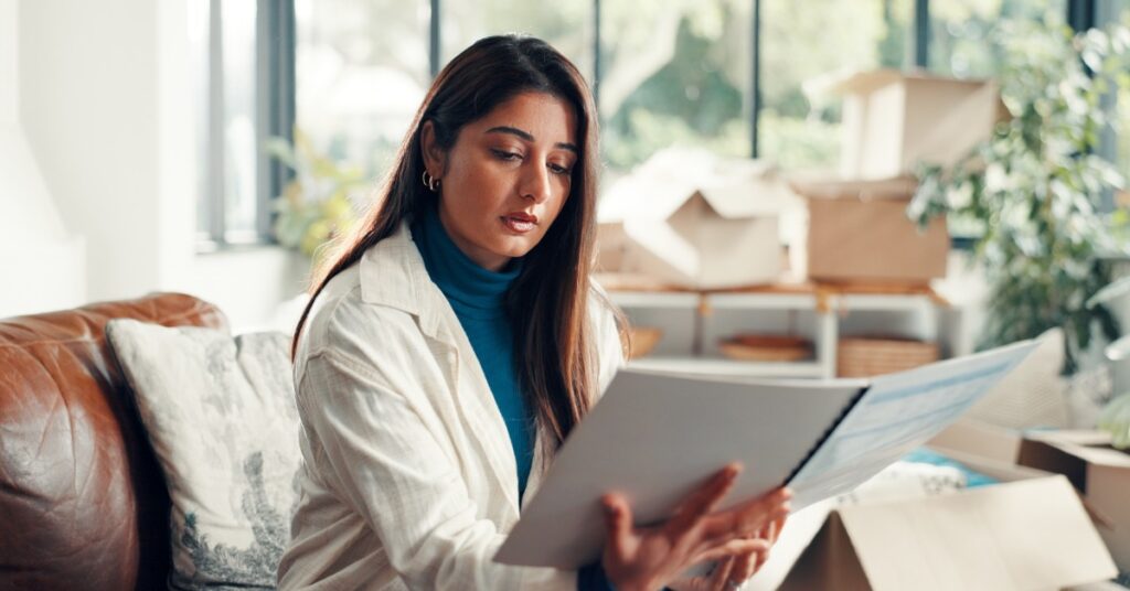 A woman wearing a blue turtleneck and a white blouse sitting on a couch in a living room while reading a document.