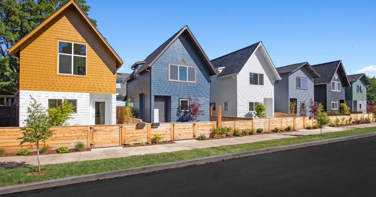 Six homes with barn-style roofs and different colored facades placed next to each other, divided by a wooden fence.