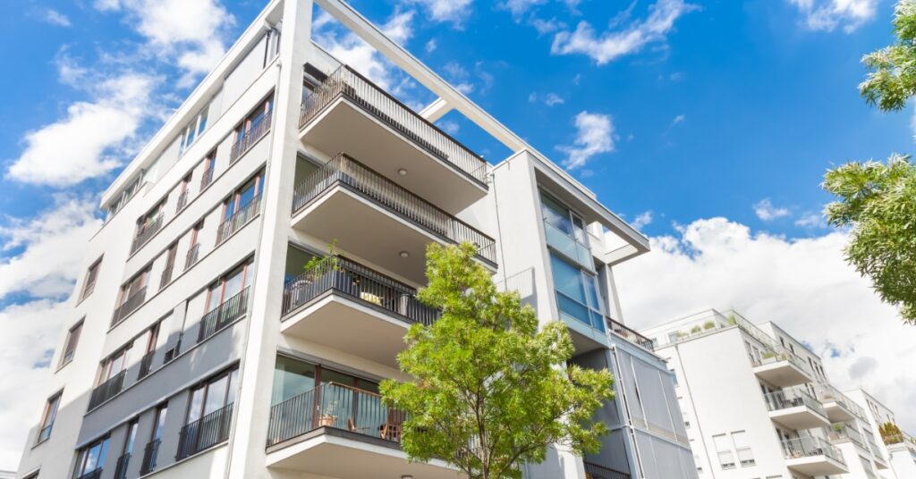 A white building with multiple apartments and small balconies, next to another similar building surrounded by trees.