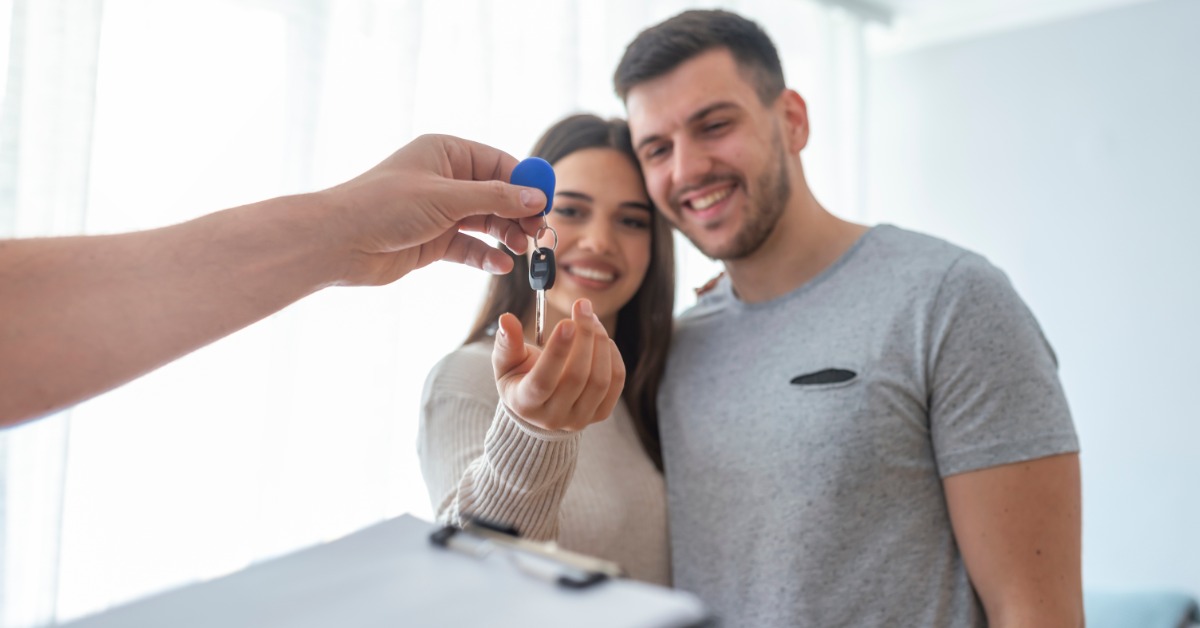 A close-up of a hand handing a key with a blue keychain to a woman standing next to a man inside a room.