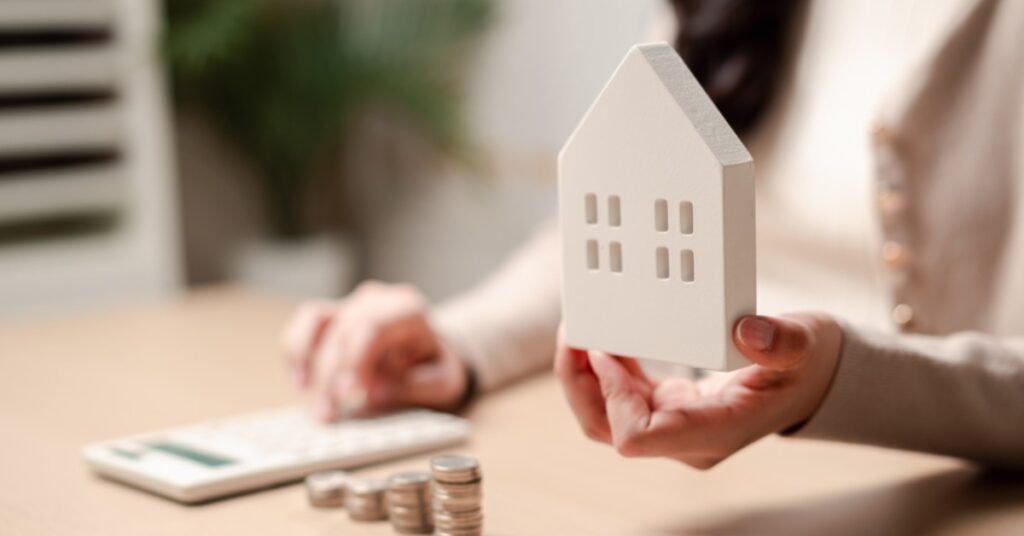 A close-up of a hand holding a house-shaped foam sitting behind a desk with stacks of coins, using a calculator.