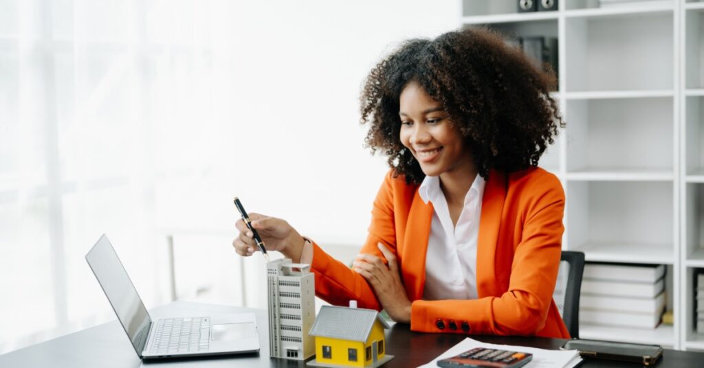 A woman with curly hair wearing a bright orange jacket behind a desk with a laptop, a model building, and a house.