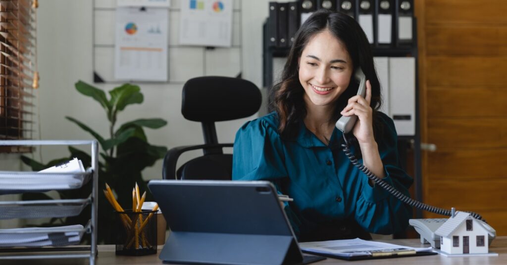 A woman on the phone looking at a tablet while sitting behind a desk in an office with multiple files in the back.