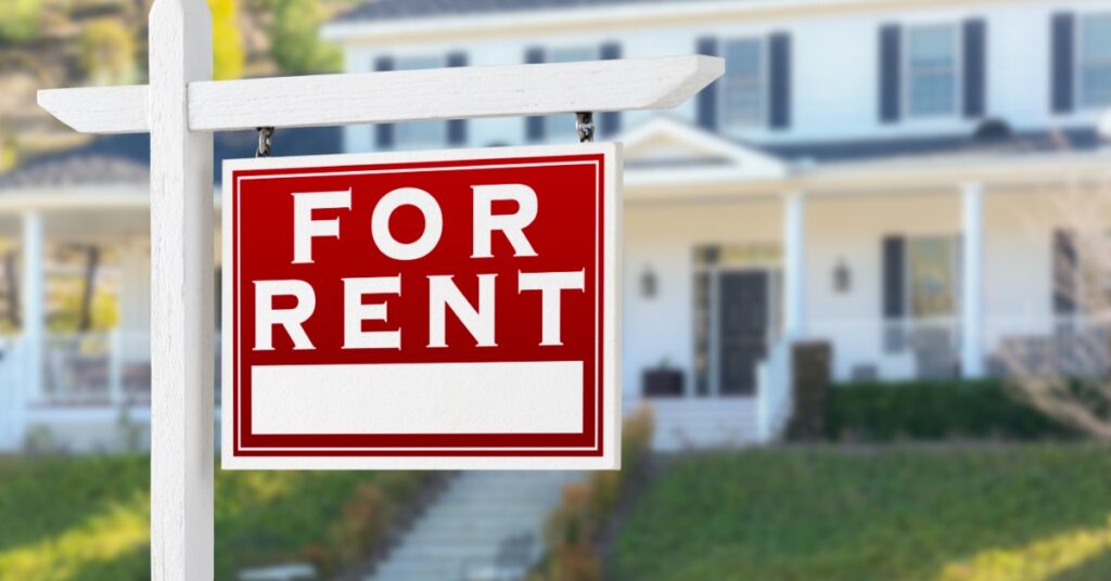 A red “For Rent” sign hanging from a white wooden structure with a blurred two-story home in the background.