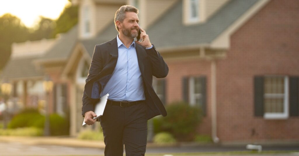 A man wearing a suit and a blue shirt walking down a street next to homes while on the phone and holding a clipboard.