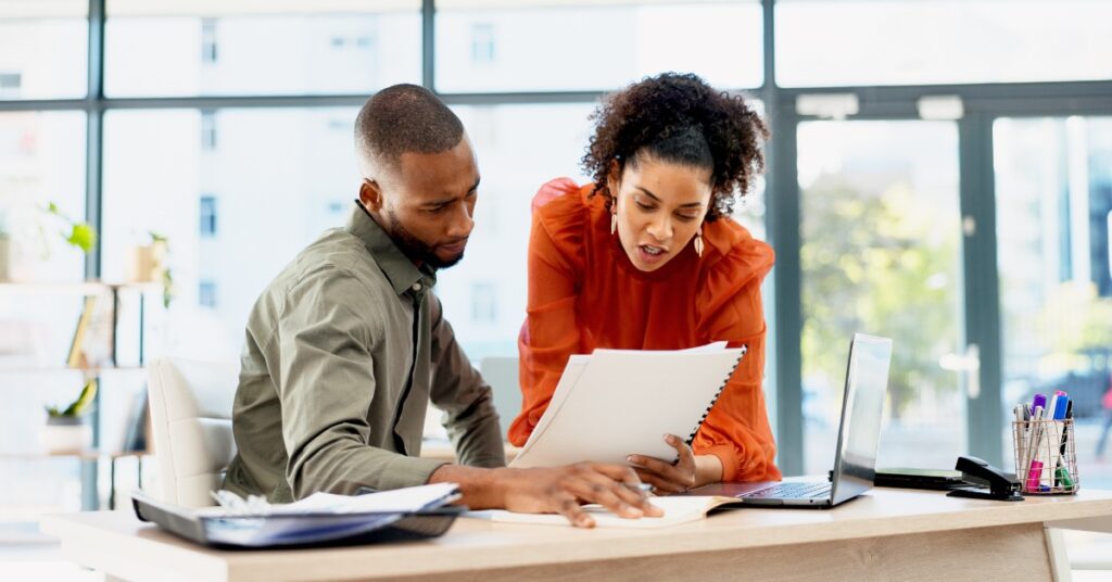 A woman wearing an orange blouse leaning on a desk next to a man sitting on a chair, both looking at a document.